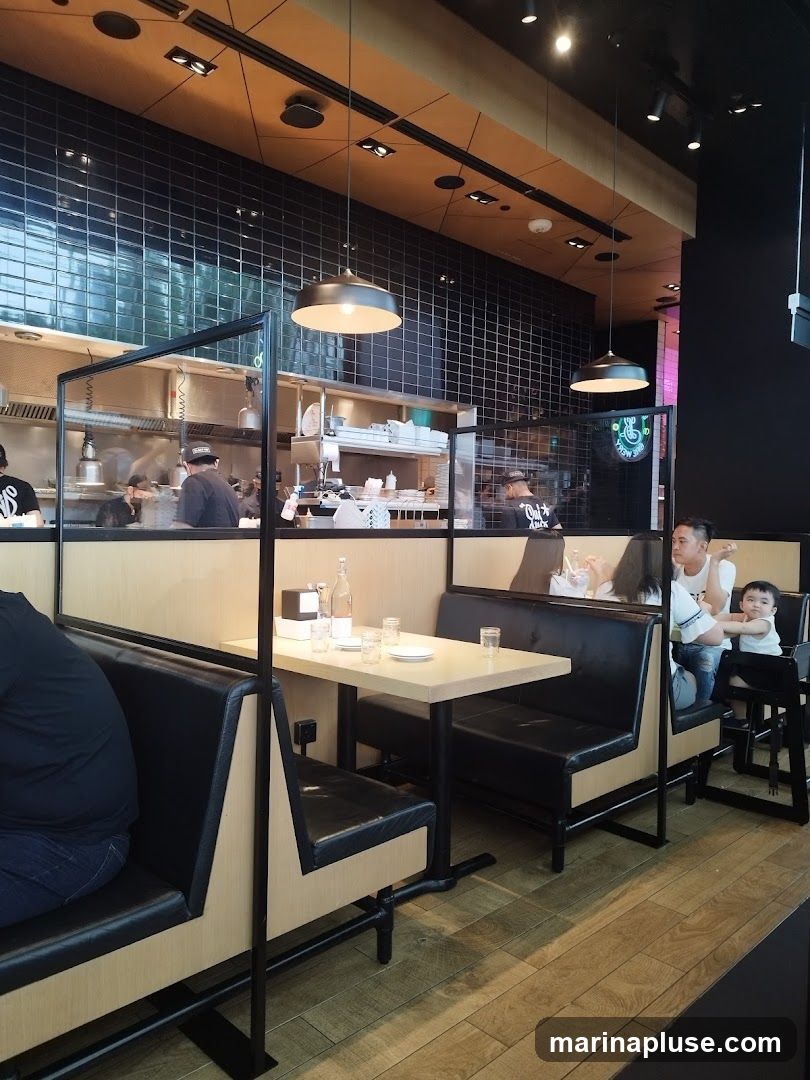 The image shows the interior of a restaurant with booth seating and a view into the kitchen. The booths are upholstered in black leather with light wood accents. Transparent dividers separate the booths. The kitchen area features stainless steel equipment and black tiled walls. Several diners are seated in the booths, including a family with a child. Pendant lights hang from the ceiling.