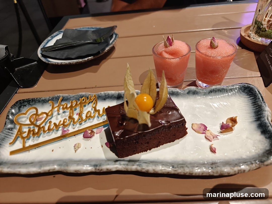 A slice of chocolate cake with a golden "Happy Anniversary" topper and a physalis fruit sits on a decorative platter. Two pink slushy drinks with rosebuds on top are in the background. Dried rose petals are scattered on the platter.