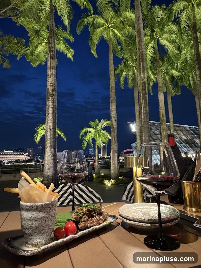 A table set for dining outdoors at night, with palm trees in the foreground and a city skyline in the background. On the table, there are two glasses of red wine, a plate of skewers with garnish, cherry tomatoes, and a cup of french fries. The ambiance is sophisticated and relaxed, with soft lighting illuminating the palm trees and the distant cityscape.