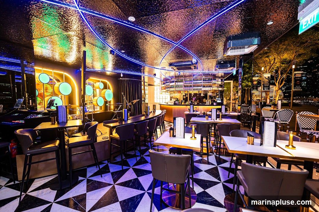 The interior of a modern bar features a black and white geometric tiled floor, a long bar area with bottles displayed on shelves behind it, and numerous tables and chairs. Blue neon lights are integrated into the ceiling, which has a textured, reflective surface. A stage area with musical equipment and circular green lights is visible on the left. The overall ambiance is chic and contemporary, with a view of city lights visible through the windows.