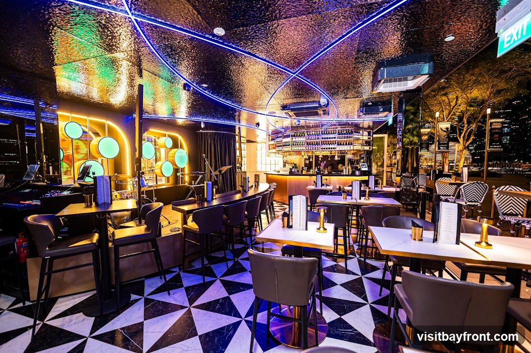 The interior of a modern bar features a black and white geometric tiled floor, a long bar area with bottles displayed on shelves behind it, and numerous tables and chairs. Blue neon lights are integrated into the ceiling, which has a textured, reflective surface. A stage area with musical equipment and circular green lights is visible on the left. The overall ambiance is chic and contemporary, with a view of city lights visible through the windows.