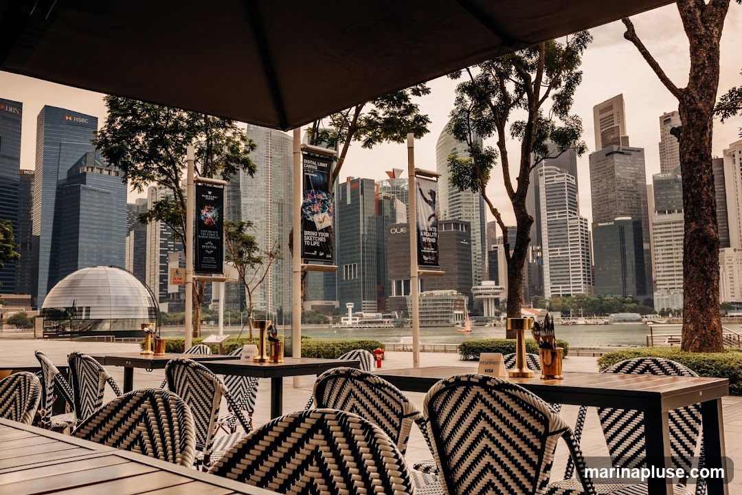 An outdoor seating area with tables and chairs is set up in front of a city skyline. The tables are set with cutlery and golden candle holders. The chairs have a black and white chevron pattern. In the background, several tall skyscrapers are visible, along with a domed building and a body of water. Banners with text and images are hanging from poles in front of the buildings.
