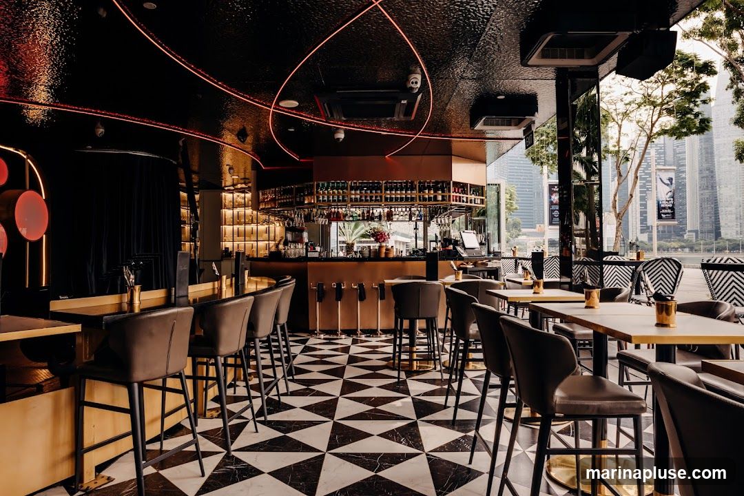 The interior of a stylish bar with a black and gold color scheme. The floor features a black and white geometric pattern. A long bar with stools is visible, stocked with bottles of alcohol. Tables and chairs are arranged throughout the space, some with a view of the city skyline outside. Red neon lighting accents the ceiling.