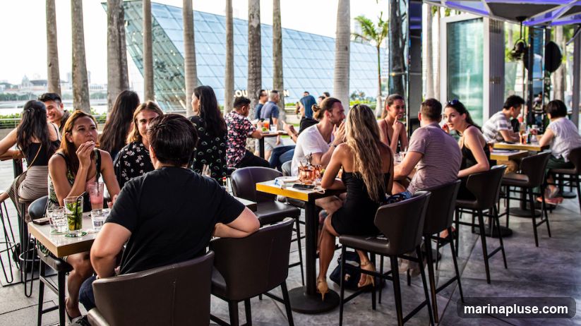 A group of people are seated at tables in an outdoor dining area. The background features a modern building with a glass facade and palm trees. The patrons are dressed casually and appear to be enjoying drinks and conversation.