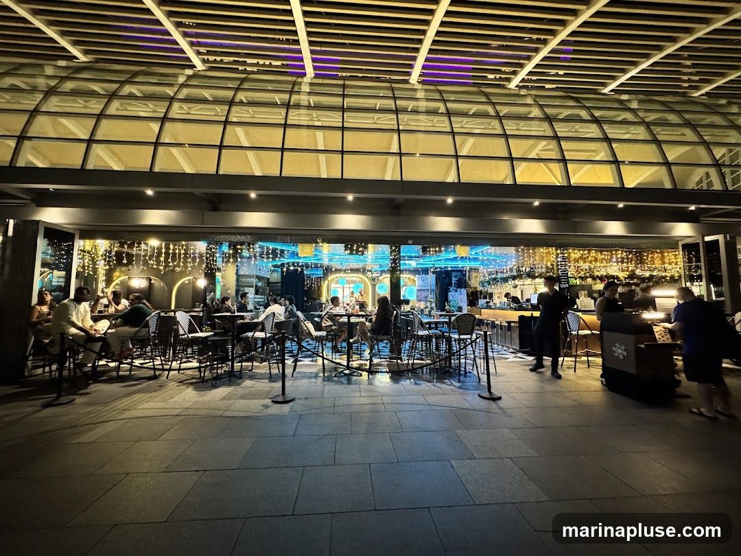 An outdoor seating area of a restaurant at night. Tables and chairs are set up on a paved patio. Inside the restaurant, people are seated at tables and at the bar. String lights are hung across the ceiling and walls, creating a festive atmosphere.