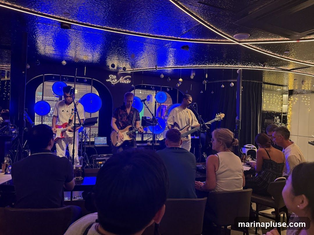 A live band performs on a stage in a dimly lit venue. The band consists of three musicians playing guitars and a bass. The stage is decorated with blue circular lights and hanging ornaments. Several patrons are seated at tables, watching the performance. The venue has a modern interior with a textured ceiling and neon lighting. A sign above the stage reads "Le Nola".