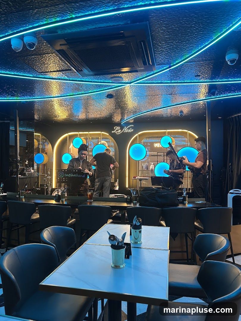 A dimly lit interior of a venue with a live band performing on a stage. The stage area is illuminated by blue circular lights and neon blue and green lighting accents the ceiling. Tables and chairs are set up in the foreground, with some set for dining. The band members are visible playing instruments. The venue appears to be a bar or restaurant.