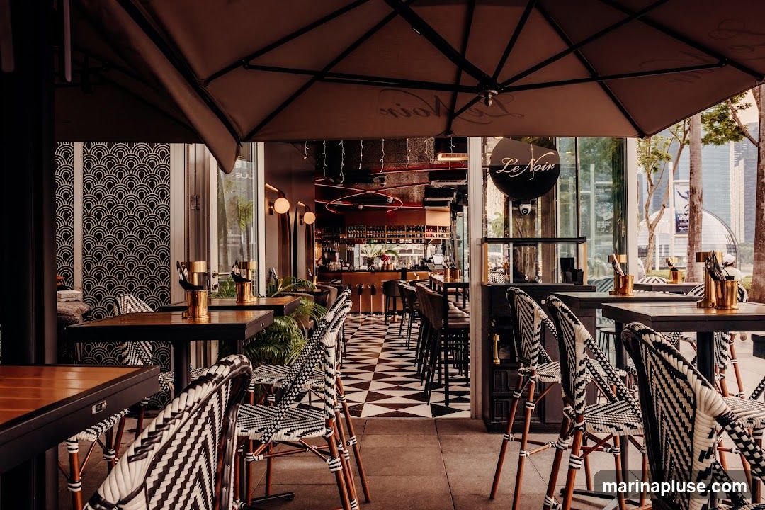 An outdoor seating area of a restaurant with tables and chairs. The interior of the restaurant is visible in the background, featuring a bar and seating. The restaurant's name, 'Le Noir', is displayed on a circular sign.
