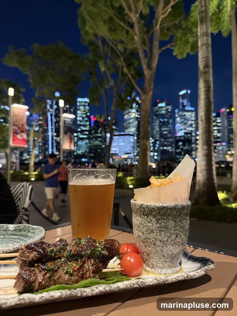 A close-up of a meal served outdoors at night, featuring grilled meat skewers with a green sauce, cherry tomatoes, a glass of beer, and a small portion of fries. In the background, a blurred cityscape with illuminated skyscrapers is visible, along with trees and palm trees.