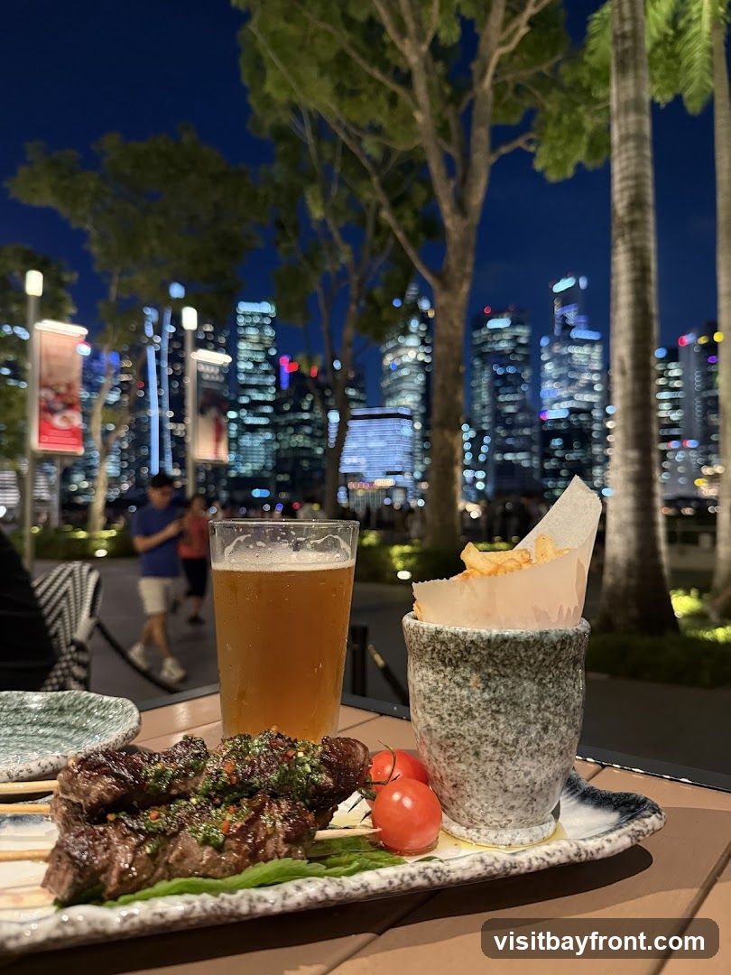 A close-up of a meal served outdoors at night, featuring grilled meat skewers with a green sauce, cherry tomatoes, a glass of beer, and a small portion of fries. In the background, a blurred cityscape with illuminated skyscrapers is visible, along with trees and palm trees.