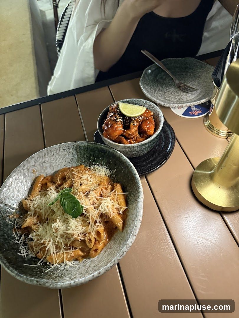 A bowl of penne pasta with red sauce and grated cheese is served on a table, next to a small bowl of glazed chicken bites topped with a lime wedge. A person is seated at the table, partially visible. A coaster with a Tiger logo is also visible.