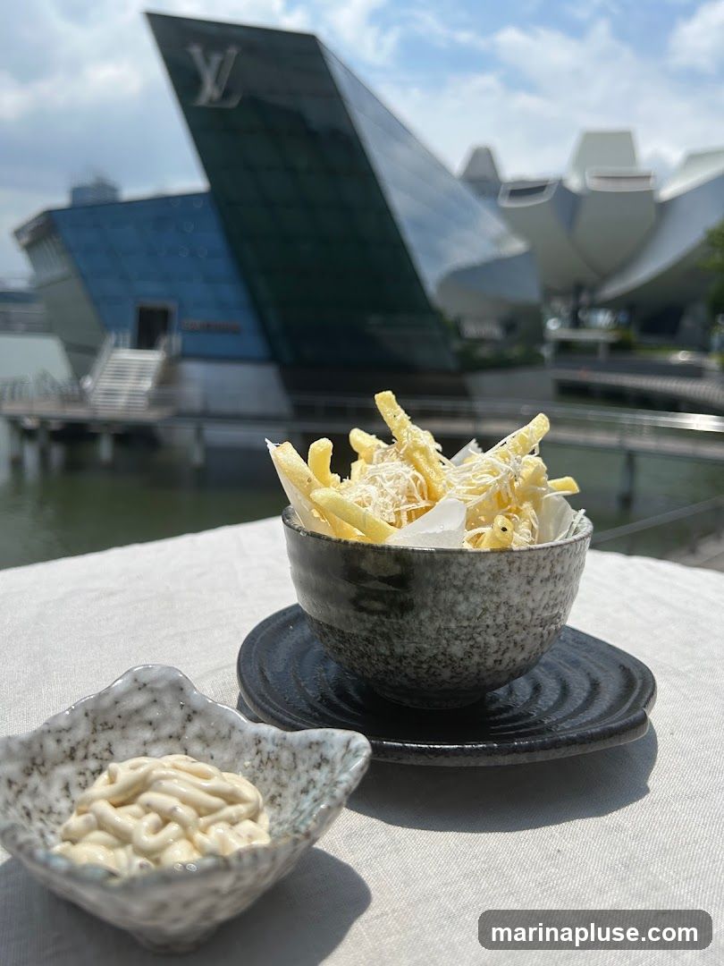 A close-up shot of truffle fries served in a dark speckled bowl, topped with grated cheese. Beside it, a small dish holds a dollop of aioli. In the background, a modern architectural building with a prominent Louis Vuitton logo is visible under a bright sky. The setting appears to be outdoors, possibly a restaurant patio.