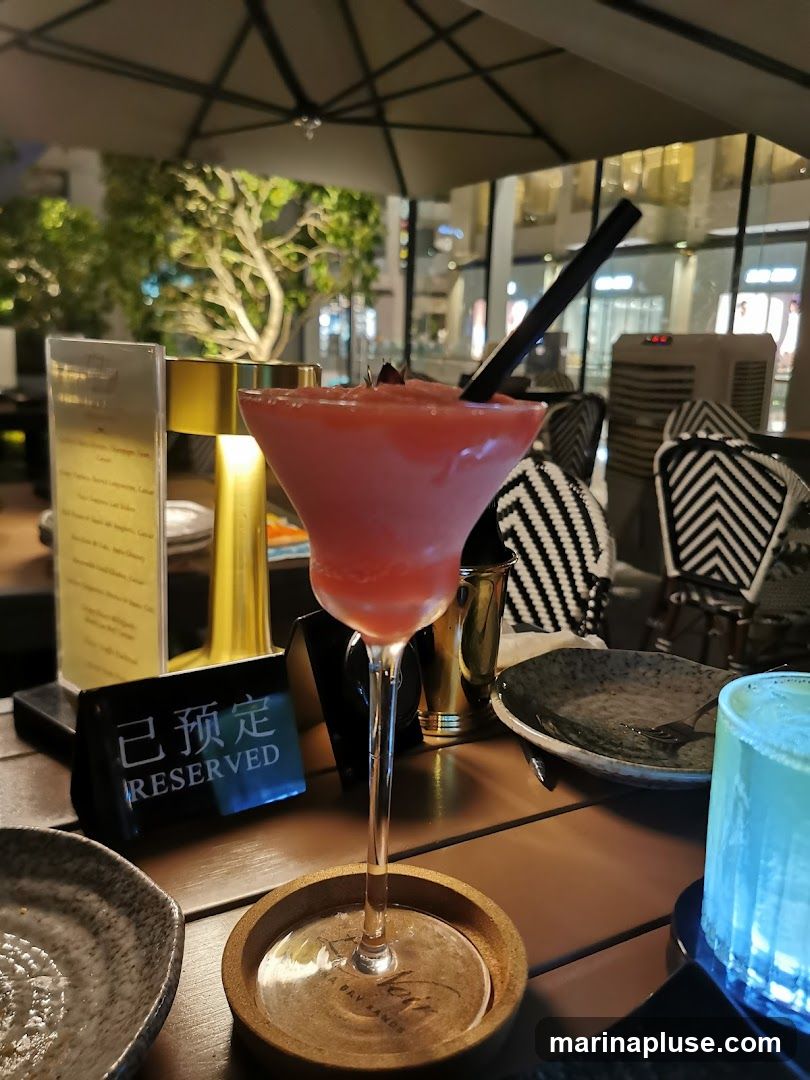 A table at an outdoor seating area with a pink frozen cocktail in a martini glass. A black straw is in the drink. A coaster with the text "Veri A BAY LANDS" is under the glass. A black sign with "RESERVED" in white text is on the table. There are plates, cutlery, and a blue glowing glass on the table. In the background, there are chairs with black and white patterned upholstery, trees, and buildings.