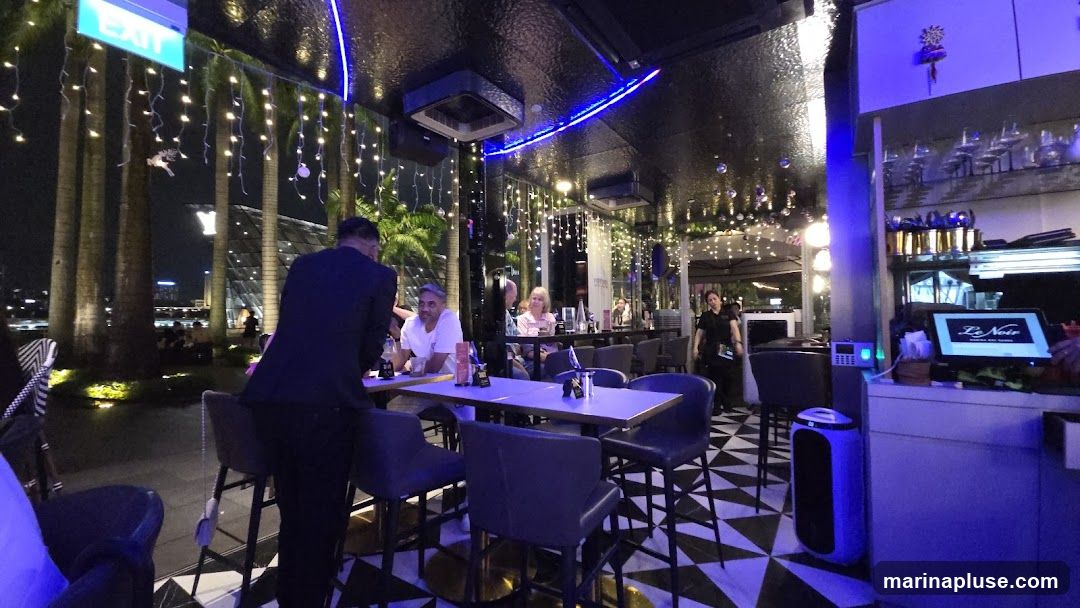 An interior view of a bar or restaurant at night. Palm trees and string lights are visible outside. Several patrons are seated at tables, and a server is standing near a bar area. A POS system with the logo "Le Noir" is visible on the counter.