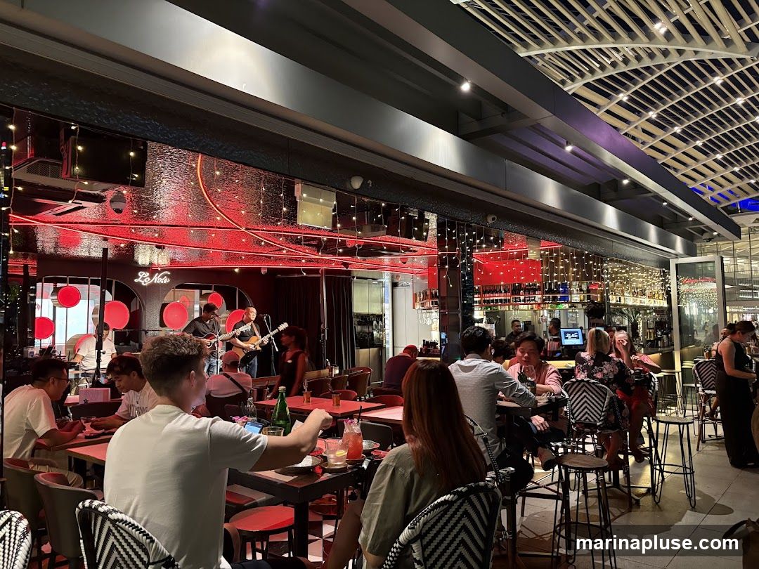 A lively restaurant interior with patrons seated at tables, enjoying drinks and a live band performing on a stage. The decor features red accents, string lights, and mirrored walls.