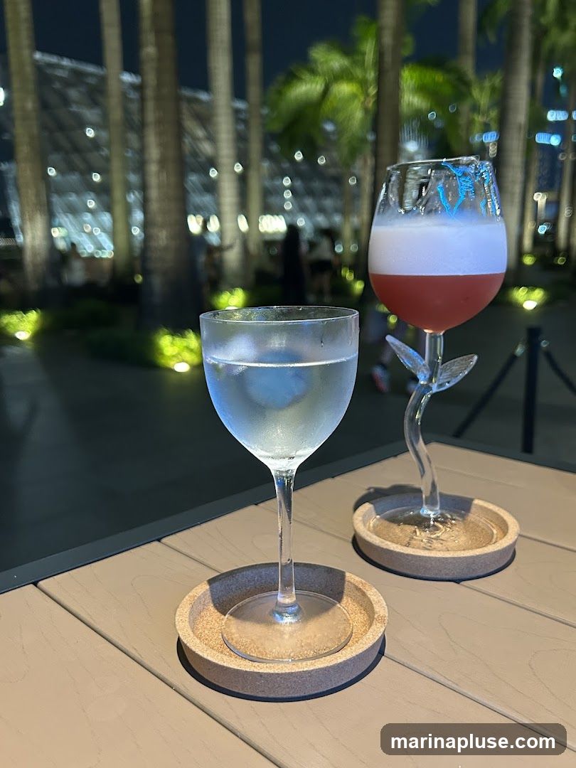 Two cocktails are placed on a wooden table outdoors at night. The drink in the foreground is in a stemmed glass and appears to be clear with ice. The drink in the background is in a unique stemmed glass shaped like a flower stem and has a pinkish-red liquid topped with white foam. In the background, palm trees and illuminated structures are visible, suggesting a tropical or resort setting.