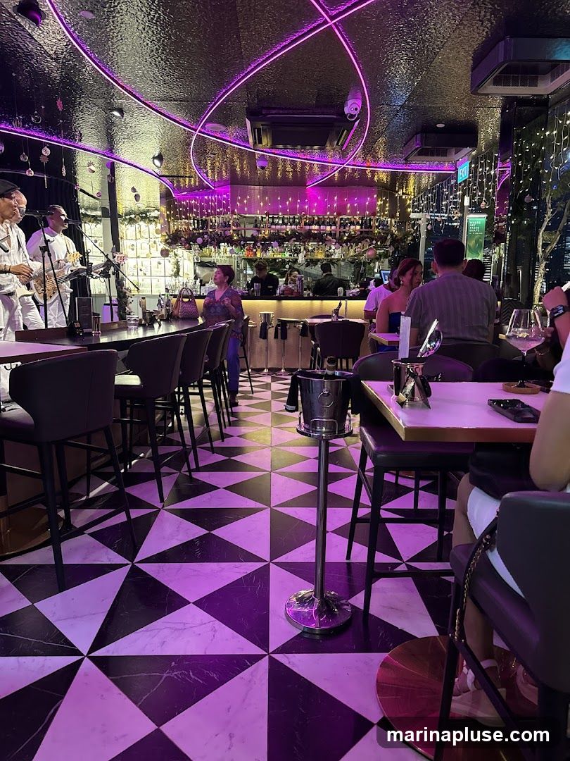 The interior of a bar with a live band playing on a stage. The room features a black and white geometric tiled floor, a long bar with stools, and a well-stocked liquor shelf behind the bar. Purple neon lights illuminate the ceiling and walls. Several patrons are seated at tables and the bar.
