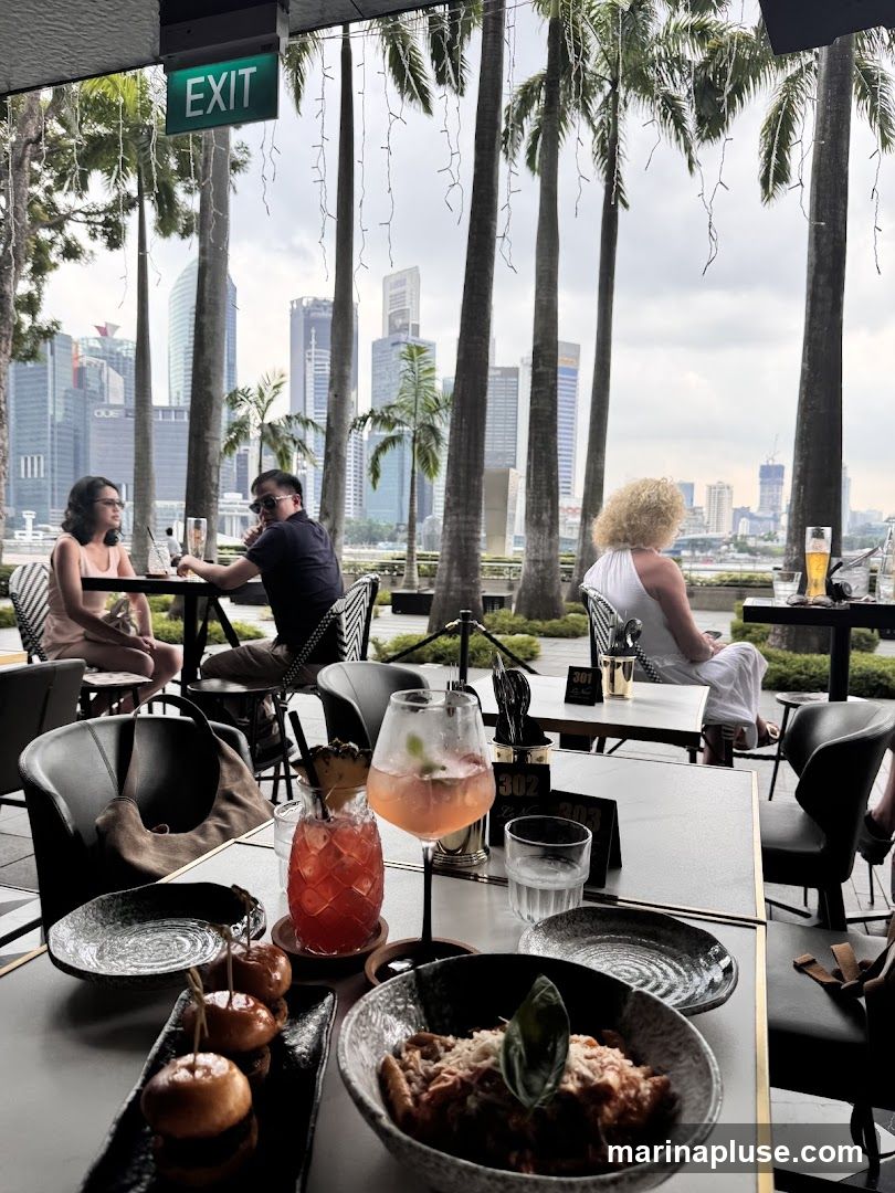 An outdoor dining area with tables set for meals, overlooking a city skyline with tall buildings and palm trees. Several people are seated at tables, enjoying drinks and food. Two colorful cocktails are prominently displayed on a table in the foreground, along with a dish of pasta and small buns. An "EXIT" sign is visible overhead.