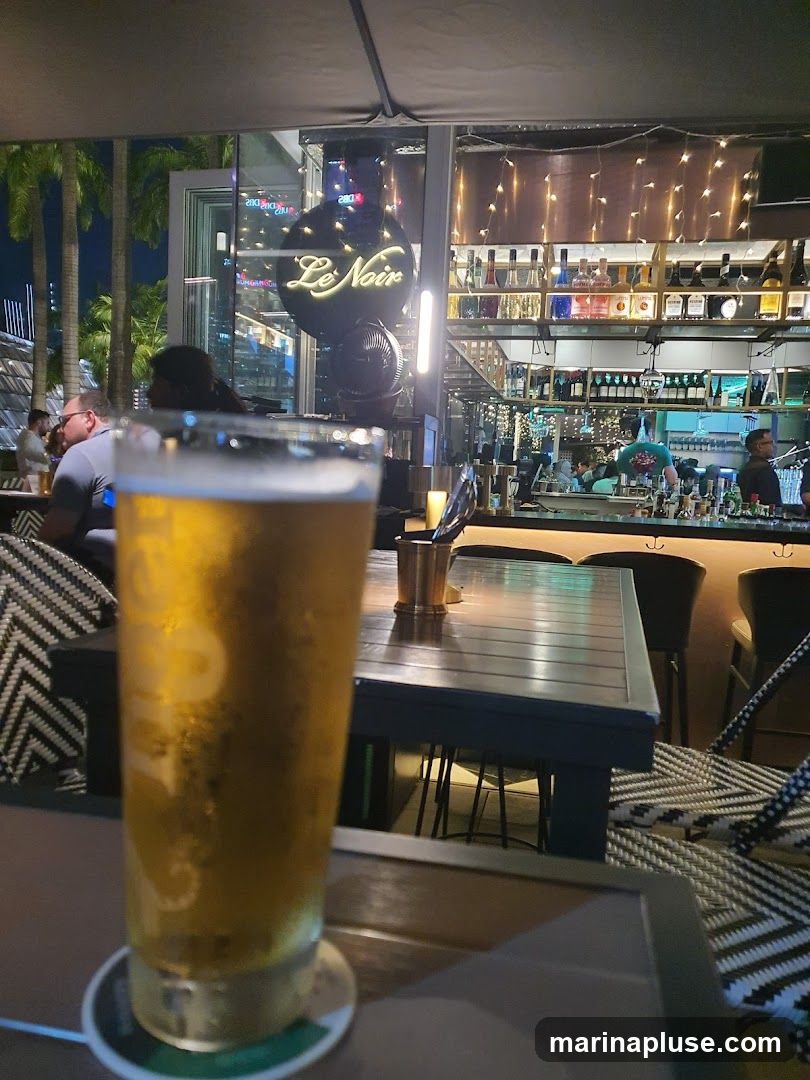 A glass of beer is in the foreground, with a bar and seating area in the background. The bar features shelves stocked with bottles and decorative lights. The sign 'Le Noir' is visible.