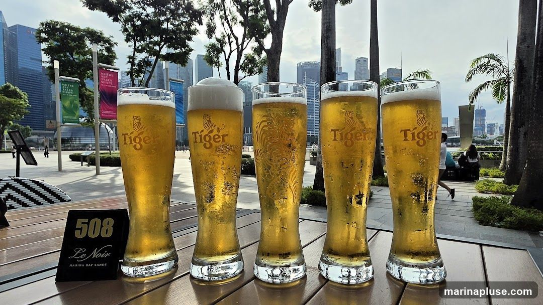 Five tall glasses of Tiger beer are lined up on a wooden table. The glasses are condensation-covered and filled with golden beer, topped with foam. The Tiger logo is visible on most of the glasses. In the background, a cityscape with tall buildings and palm trees can be seen. A table number sign reads "508 Le Noir MARINA BAY SANDS".