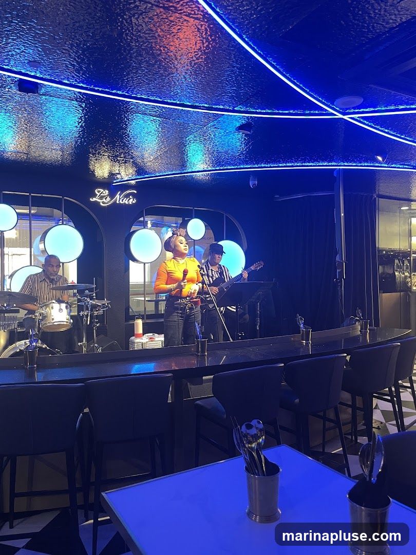 A live band performs on a stage in a dimly lit venue with blue neon lighting. A female singer is at the microphone, with a drummer to her left and a guitarist to her right. The stage has a sign that reads "Le Nuis". In the foreground, there are bar stools and tables with cutlery holders.