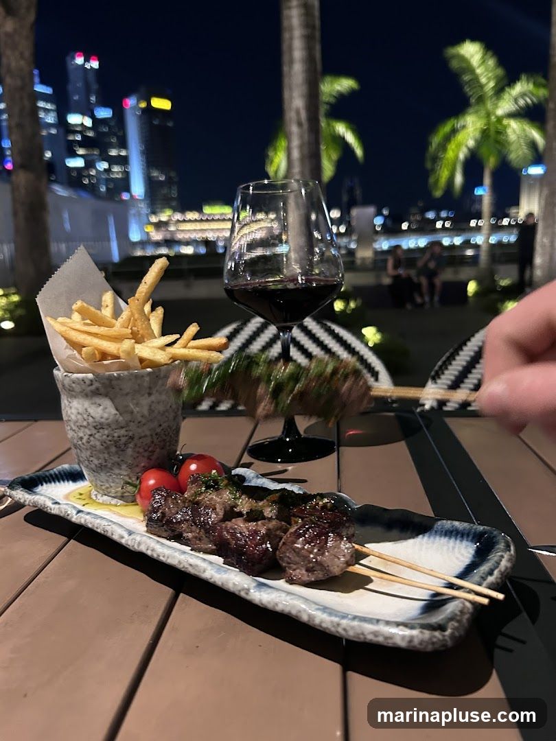 A plate of grilled meat skewers and a side of french fries are served on a table at night. A glass of red wine sits behind the food. In the background, a city skyline with illuminated skyscrapers and palm trees can be seen.