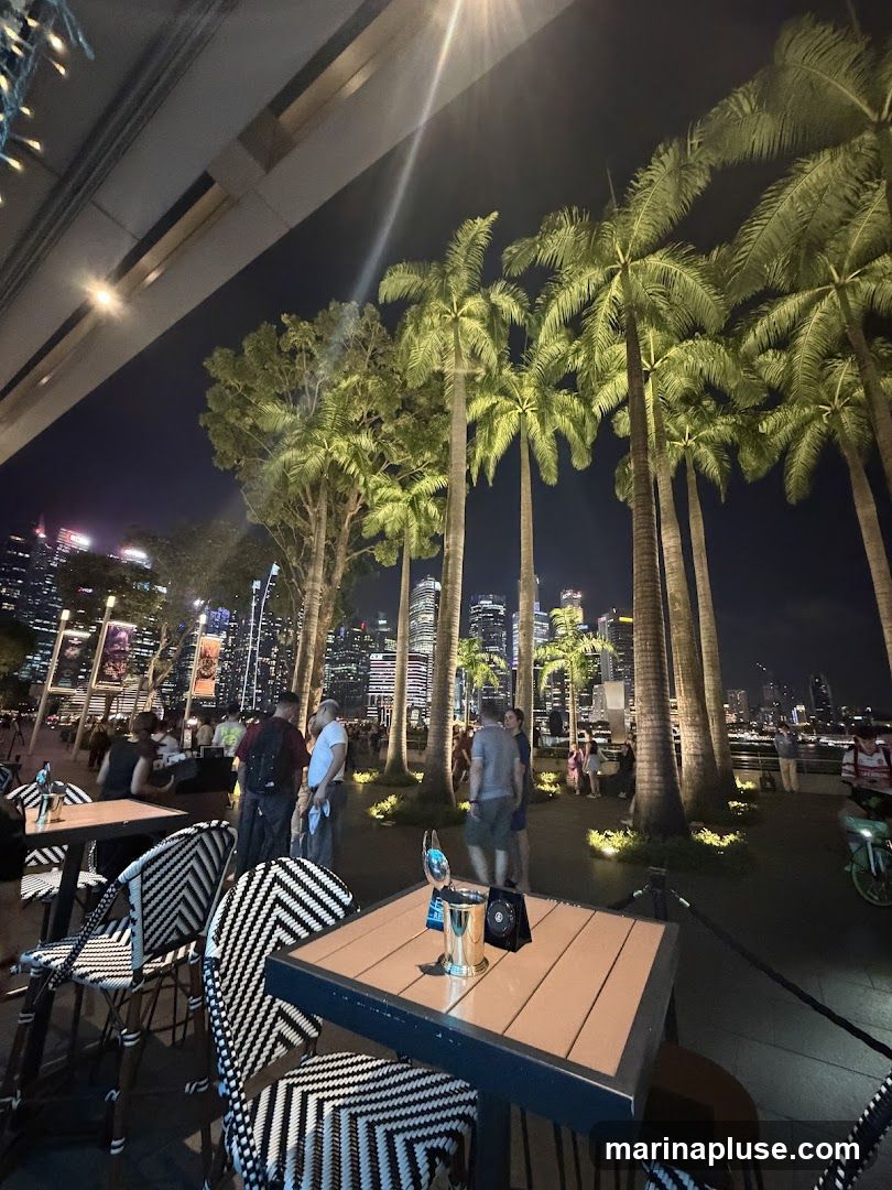 An outdoor dining area at night with tables and chairs set up. Palm trees are visible in the foreground, and a city skyline with illuminated skyscrapers is in the background. Several people are seen walking around and dining.
