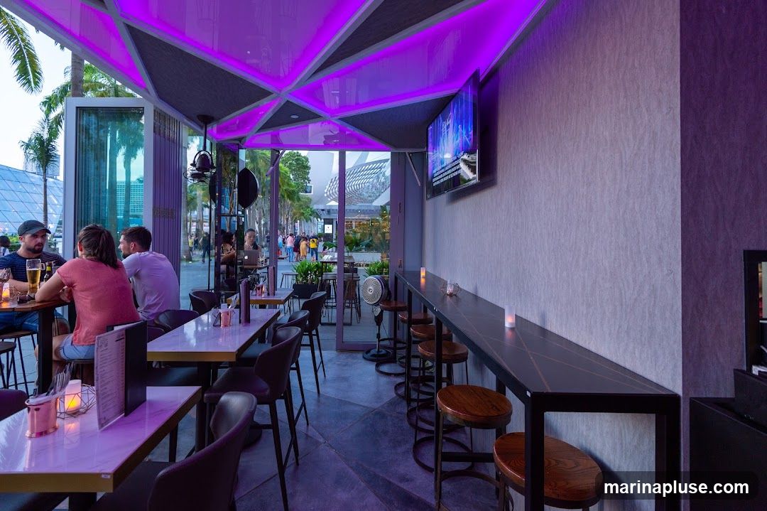 An interior view of a modern bar or restaurant with purple ambient lighting. Several tables and chairs are set up, with patrons seated at one table. A long bar counter with stools lines the wall, and a television screen displays content. The outdoor area with palm trees and people walking by is visible through the open facade.