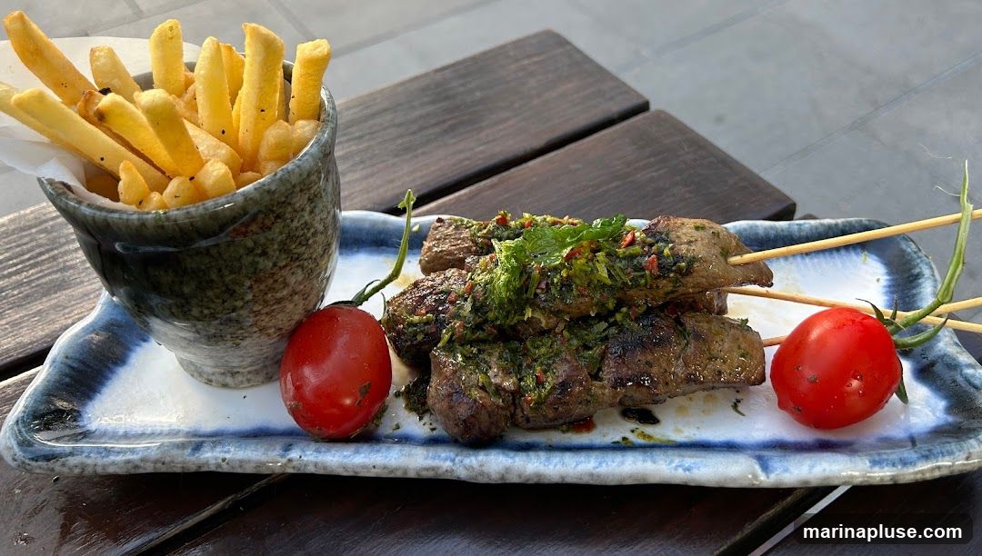 Three skewers of grilled meat topped with a green herb sauce are served on a rectangular plate with a blue and white pattern. Two whole cherry tomatoes are placed on the plate. A small bowl filled with french fries is positioned to the left of the skewers. The food is presented on a wooden table.