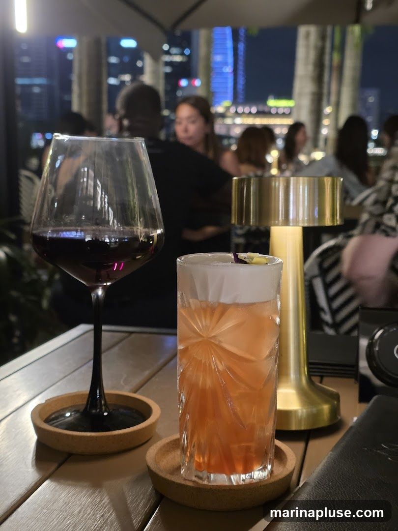 A close-up view of a table at a restaurant or bar, with two drinks and a wine glass. In the background, a blurred cityscape at night is visible, with illuminated buildings. The ambiance suggests an evening outing.