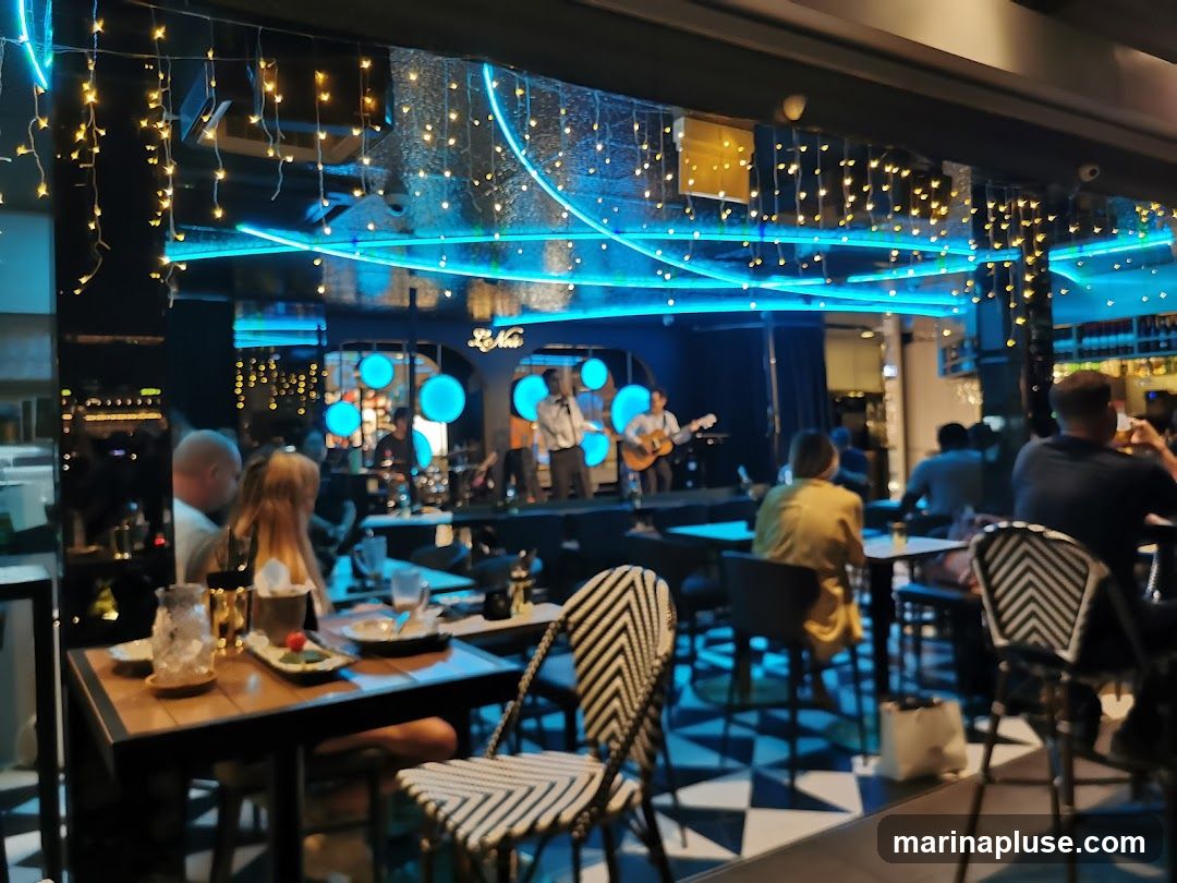 A live band performs on a stage in a dimly lit restaurant. The stage is decorated with blue neon lights and circular light fixtures. String lights hang from the ceiling, adding to the ambiance. Patrons are seated at tables, enjoying the music and drinks. The interior features a black and white checkered floor and modern seating.