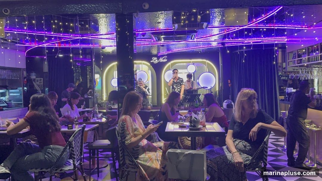 A band performs on a stage in a dimly lit restaurant with purple neon lighting and string lights. Several patrons are seated at tables, some looking at their phones, while others converse. A bartender can be seen in the background. The restaurant appears to have a modern and chic ambiance.