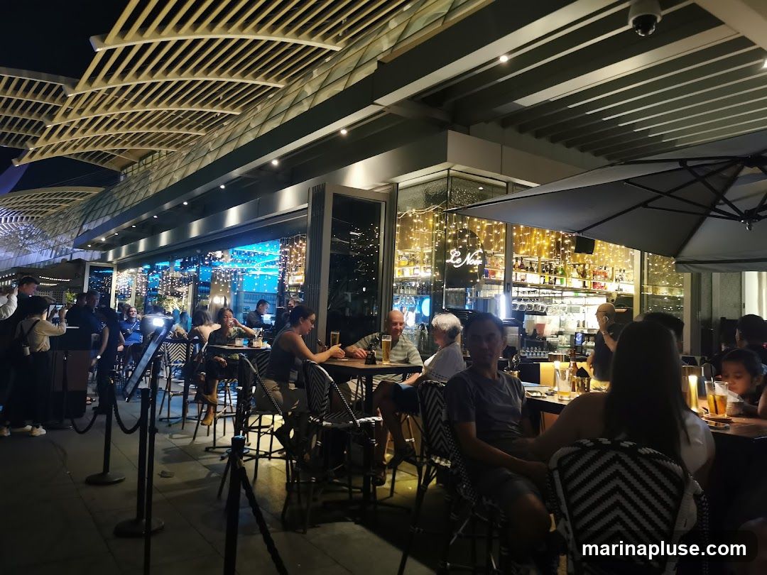 People are dining at tables outside a restaurant at night. The restaurant has a modern architectural design with a slatted roof structure. Inside, the bar area is visible with shelves stocked with bottles and illuminated by warm string lights. Several patrons are seated at tables, some with drinks and food.
