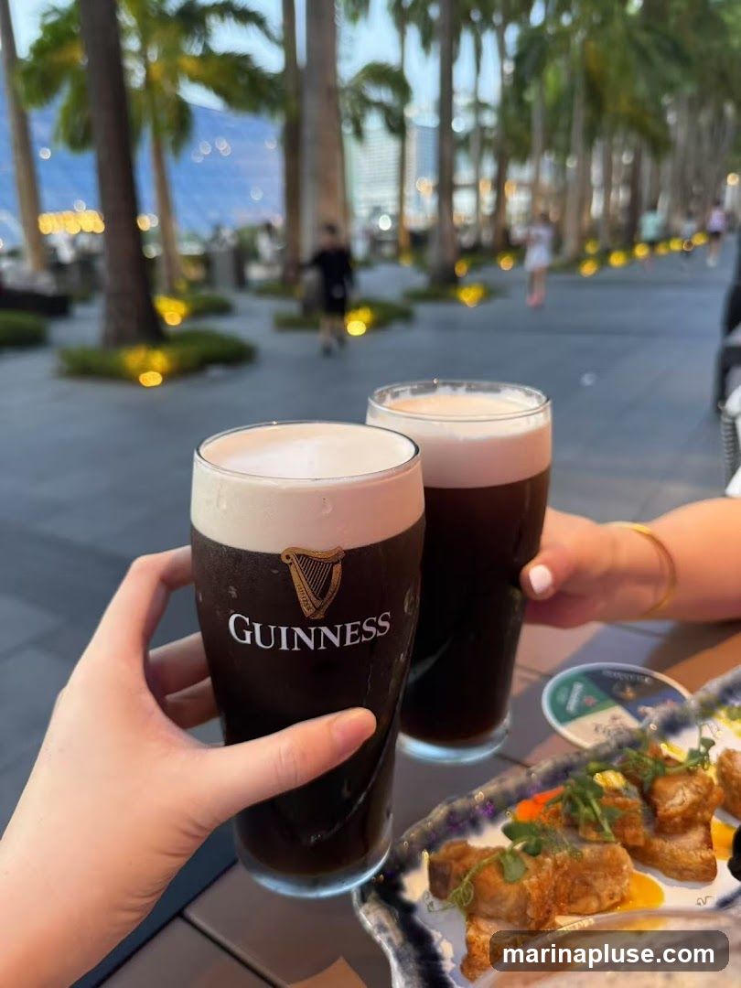 Two hands hold up glasses of Guinness beer, with a plate of fried food in the foreground. The background shows a walkway lined with palm trees and blurred figures of people.