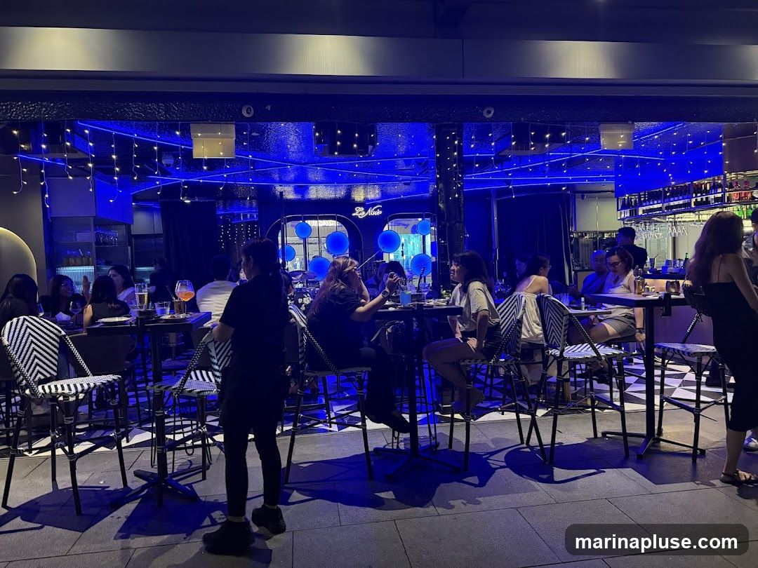 A group of people are seated at tables inside a restaurant, illuminated by vibrant blue lighting and string lights. The restaurant appears to be a bar or lounge, with a bar area visible in the background stocked with bottles and glassware. Patrons are engaged in conversation and enjoying drinks. The seating includes high-top tables with chairs featuring a black and white patterned upholstery. The overall ambiance is lively and modern.