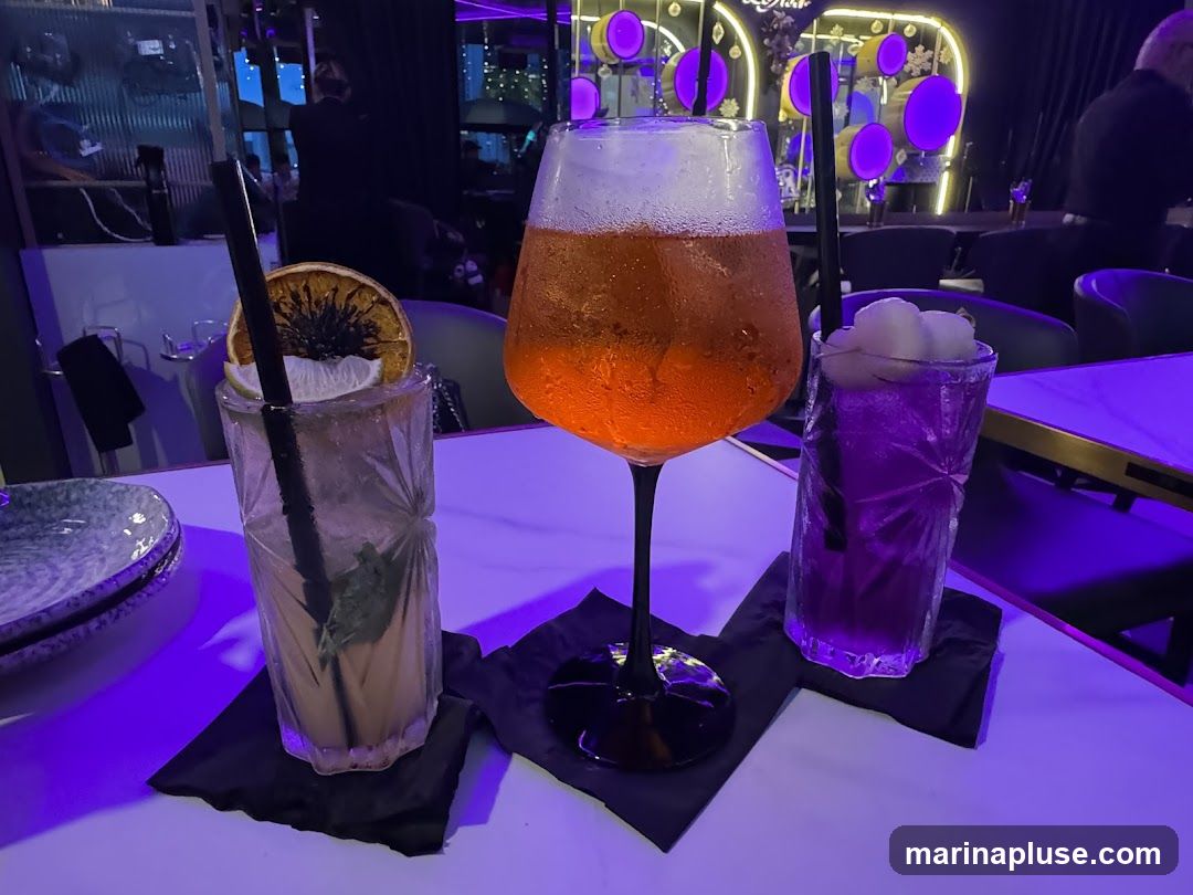 Three colorful cocktails are displayed on a white bar. The cocktail on the left is a light brown color with a dried orange slice and a lime wedge as garnish. The middle cocktail is a large glass filled with an orange-colored drink, topped with foam and ice. The cocktail on the right is a vibrant purple color, garnished with what appears to be lychee fruit. The bar area is dimly lit with purple ambient lighting, and in the background, there are blurred figures and decorative elements, suggesting a lively nightlife setting.