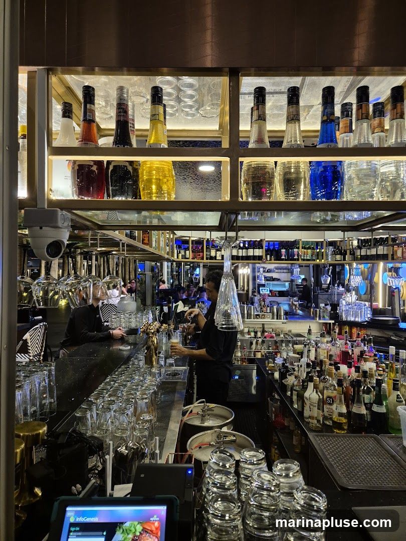 A busy bar interior with a bartender pouring a drink. Shelves behind the bar are stocked with various bottles of liquor and glassware. There are also kegs and a tablet device visible on the counter.