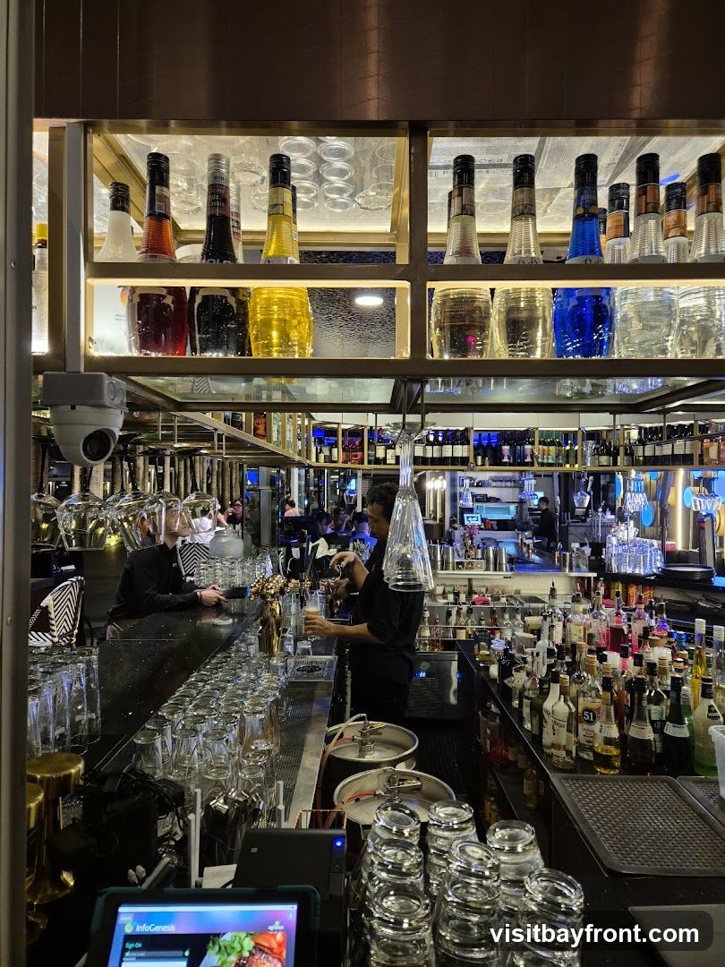 A busy bar interior with a bartender pouring a drink. Shelves behind the bar are stocked with various bottles of liquor and glassware. There are also kegs and a tablet device visible on the counter.