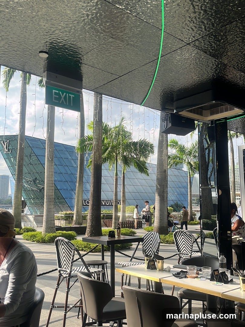 An outdoor seating area with tables and chairs is set up in front of a modern, glass-fronted building. Palm trees are scattered throughout the area, and string lights are visible. The building in the background has the text "LOUIS VUITTON" visible on its facade. People are seen walking around and sitting at tables. An "EXIT" sign is hanging from the ceiling of the seating area.