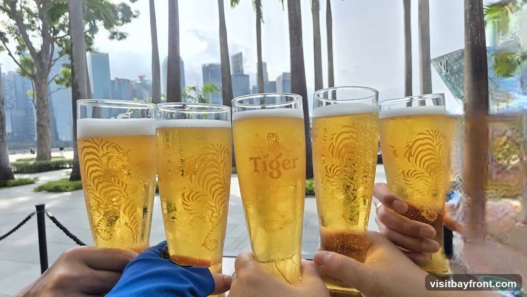 A group of people are toasting with glasses of Tiger beer. The glasses have the Tiger logo and a stylized tiger design etched into them. In the background, palm trees and modern buildings are visible, suggesting an urban outdoor setting.