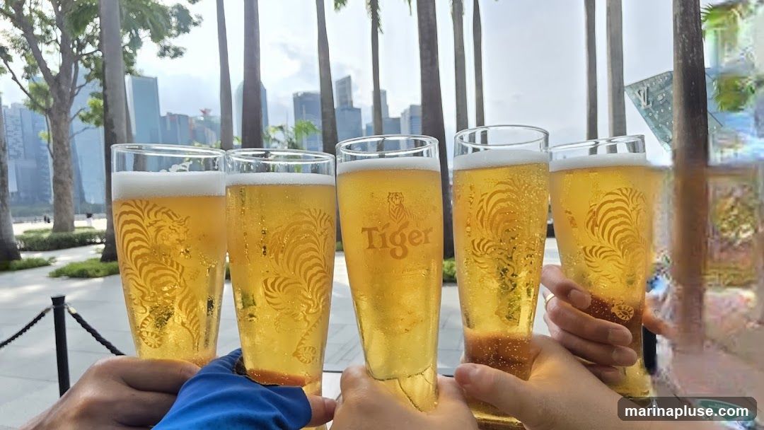 A group of people are toasting with glasses of Tiger beer. The glasses have the Tiger logo and a stylized tiger design etched into them. In the background, palm trees and modern buildings are visible, suggesting an urban outdoor setting.