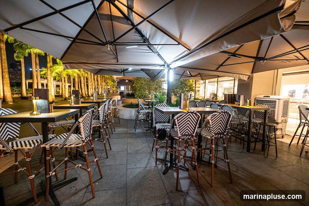 An outdoor seating area with tables and chairs is illuminated by ambient lighting under large umbrellas. Palm trees line the background, and the scene appears to be at night. The chairs have a distinctive black and white chevron pattern.