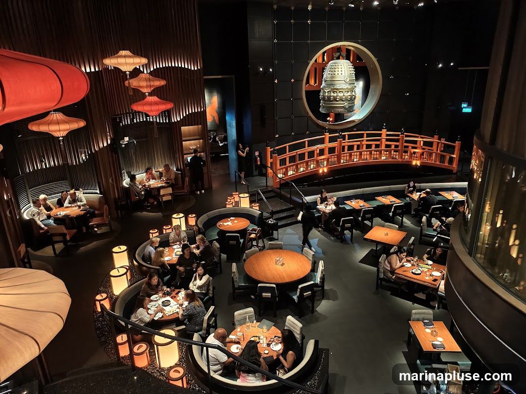 An overhead view of a dimly lit restaurant interior with a sophisticated Asian-inspired design. Several round and rectangular tables are occupied by diners. The decor features large, decorative red lanterns hanging from the ceiling, wooden paneling, and a prominent circular display with a large bell and a decorative bridge. The ambiance is elegant and intimate.