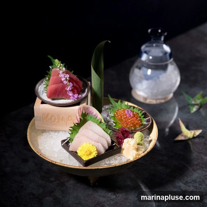 A close-up shot of a sashimi platter featuring slices of tuna and white fish, ikura (salmon roe), pickled ginger, wasabi, and garnishes. The platter is served on a bed of crushed ice in a decorative bowl. A wooden box with the word "KOMA" is visible beneath the tuna. A glass carafe filled with ice and a blue lid is in the background, along with two origami cranes.