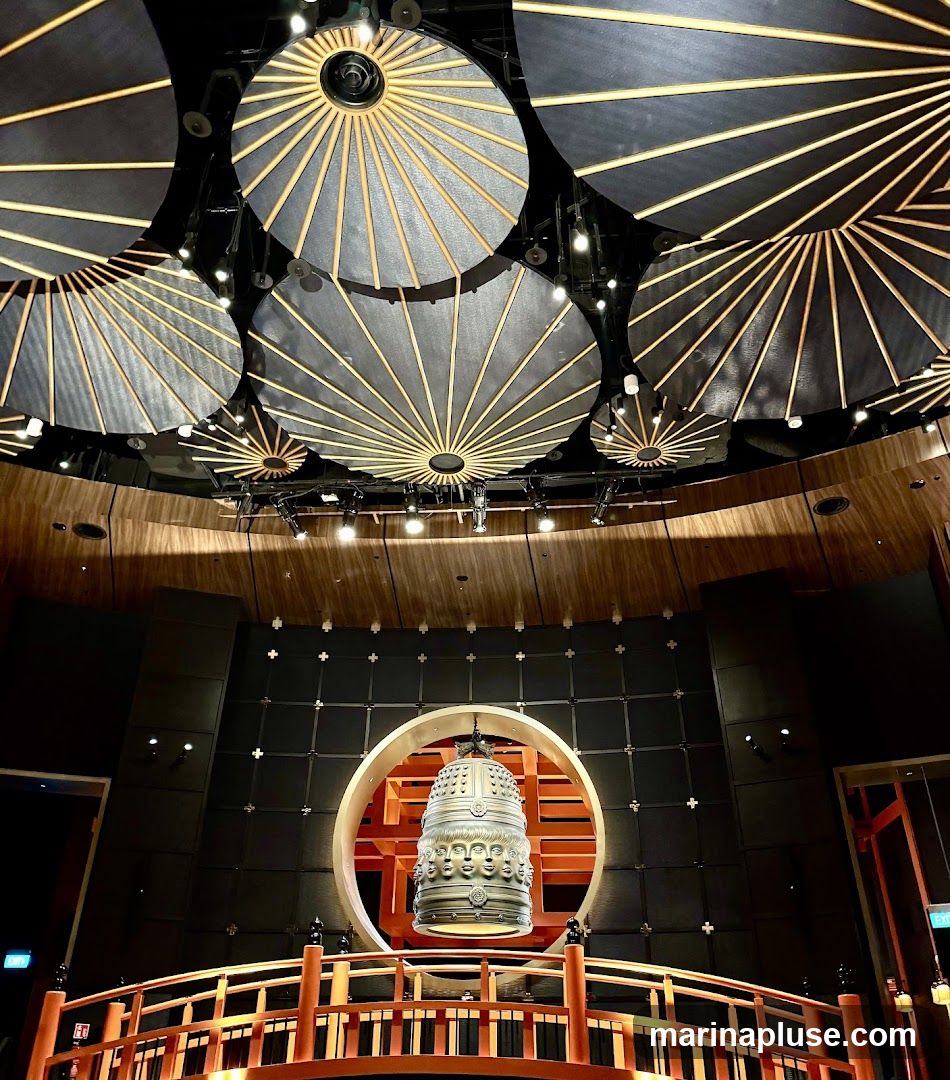 A large, ornate bell hangs within a circular opening in a dark wall. The bell is detailed with multiple faces and decorative patterns. In front of the bell is a wooden bridge with red railings. The ceiling features a series of circular, fan-like decorative elements with black fabric and wooden spokes, illuminated by spotlights. The overall ambiance is dramatic and theatrical.