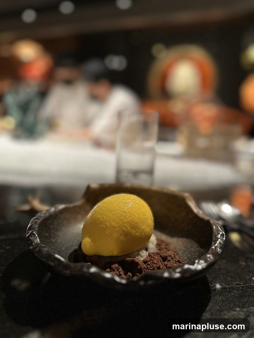 A close-up shot of a dessert featuring a scoop of bright yellow lemon sorbet served on a bed of chocolate crumble and white crystals in a dark, textured bowl. The background is blurred, showing a dimly lit restaurant interior with patrons and ambient lighting.