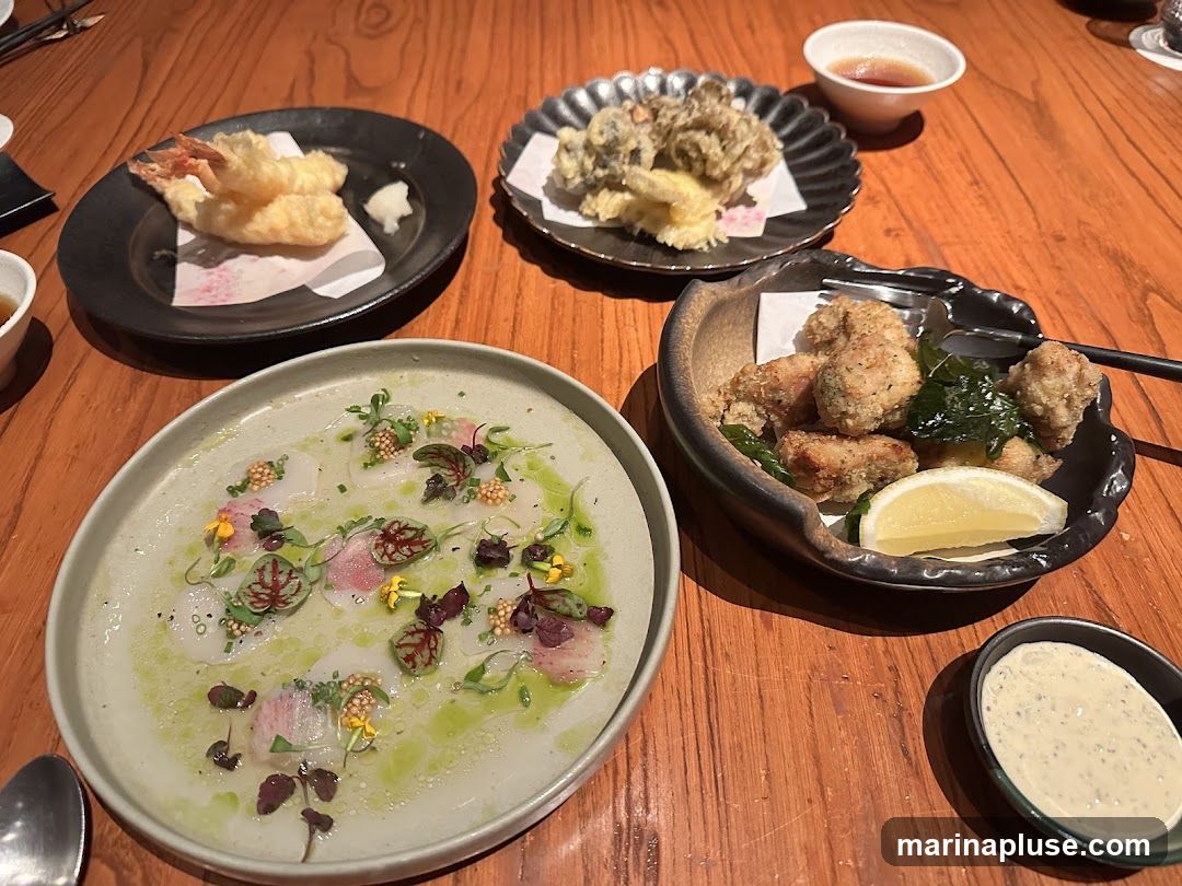 A wooden table is set with several plates of Japanese food. In the foreground, a light-colored plate holds thinly sliced raw fish, garnished with microgreens, edible flowers, and mustard seeds in a light green dressing. To the right, a dark, irregularly shaped bowl contains fried chicken pieces, a lemon wedge, and fried greens. Behind this, a black plate displays tempura-fried shrimp with a small side of grated daikon. Further back, another black plate holds a portion of mixed tempura, possibly vegetables and seafood. A small white bowl with dipping sauce is visible in the upper right corner.