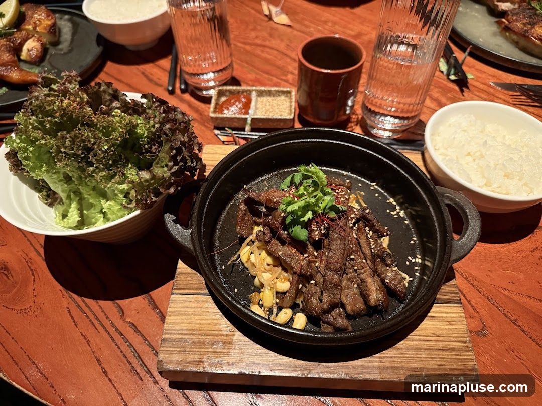 A close-up shot of a Korean BBQ meal on a wooden table. In the foreground, a cast-iron skillet holds thinly sliced grilled beef with corn and garnished with cilantro and chili threads. To the left, a bowl of fresh lettuce leaves is presented. To the right, a bowl of white rice and a glass of water are visible. In the background, another dish with grilled meat and a small dish with sauces can be seen.