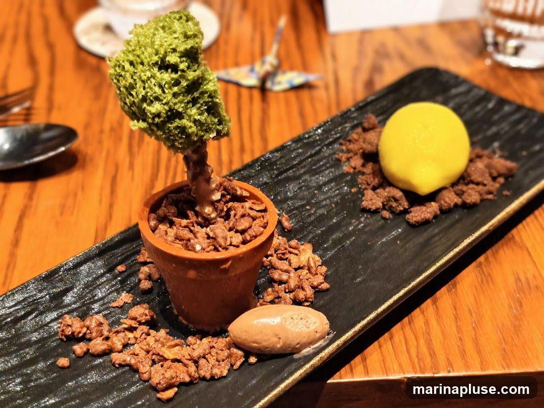A dessert is presented on a black rectangular plate. The dessert resembles a small potted tree with a green, textured top and a brown trunk, planted in a terracotta-colored pot filled with chocolate cereal. To the right of the pot, a yellow sphere, possibly a lemon sorbet or mousse, sits on a bed of chocolate crumbs. A quenelle of chocolate ice cream is placed near the bottom left of the plate. The plate is on a wooden table. In the background, a small origami crane and a glass of water are visible.
