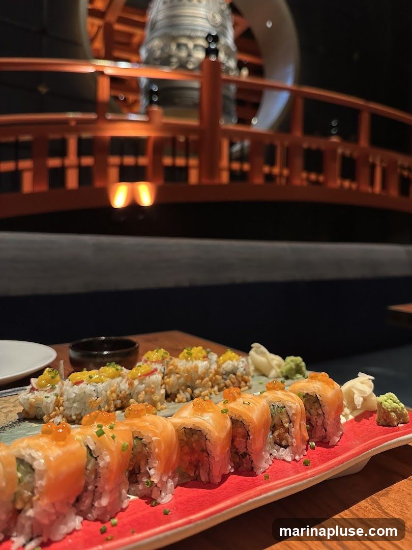 A close-up shot of a sushi platter featuring two types of sushi rolls. The front roll is topped with salmon roe and appears to be made with salmon. The roll behind it is topped with crispy rice and what looks like mango. Wasabi and pickled ginger are served alongside. In the background, a blurred view of a Japanese-inspired interior with a large bell and orange railings is visible.